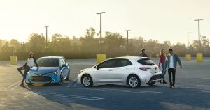Cool people hanging around 2 Corolla Hatchbacks in an empty parking lot.